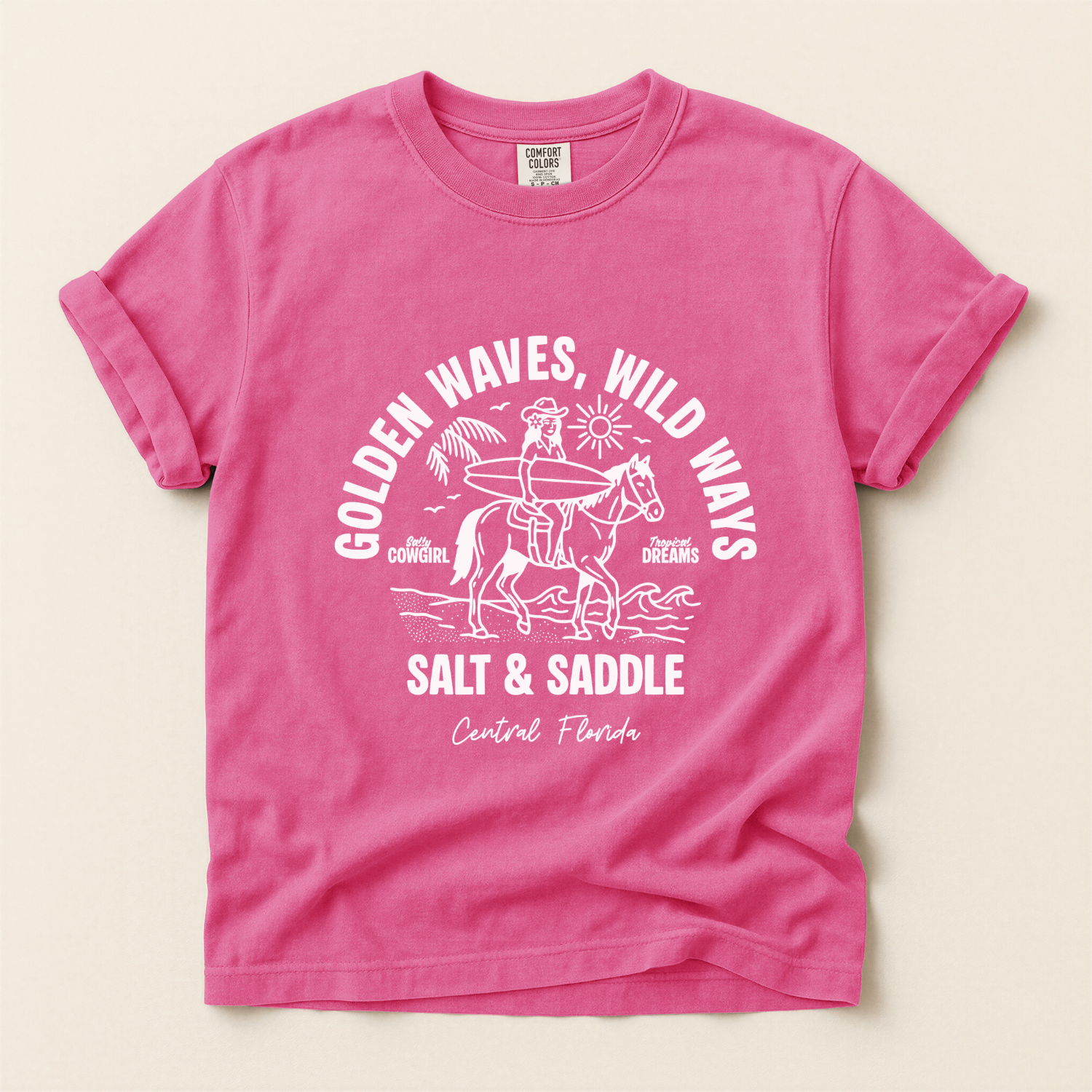 Pink t-shirt with a white graphic of a cowgirl riding a horse along the beach. The girl is holding a surfboard. The text says salt and saddle, Central Florida. Coastal Cowgirl theme.