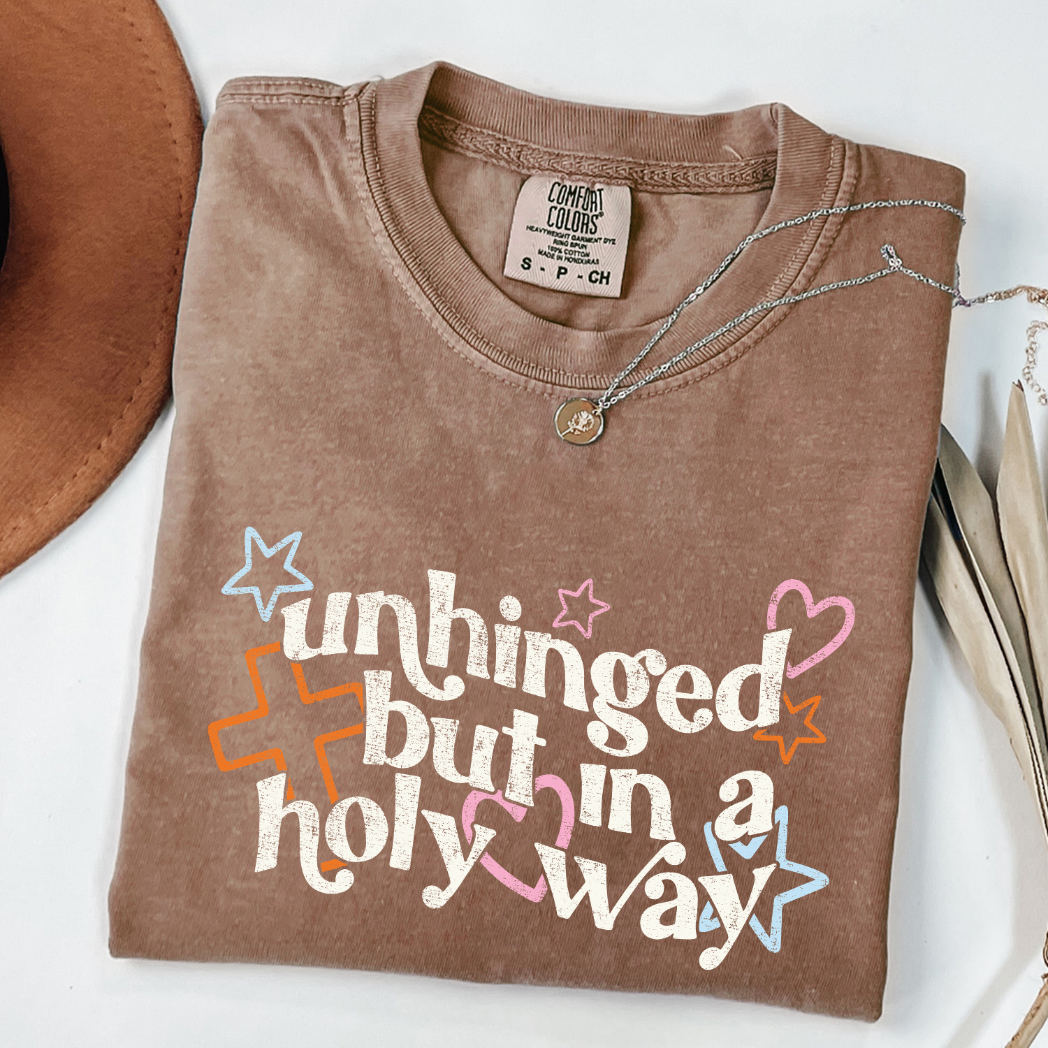 Brown t-shirt with colorful text and stars on a white surface. Text reads "unhinged but in a holy way"