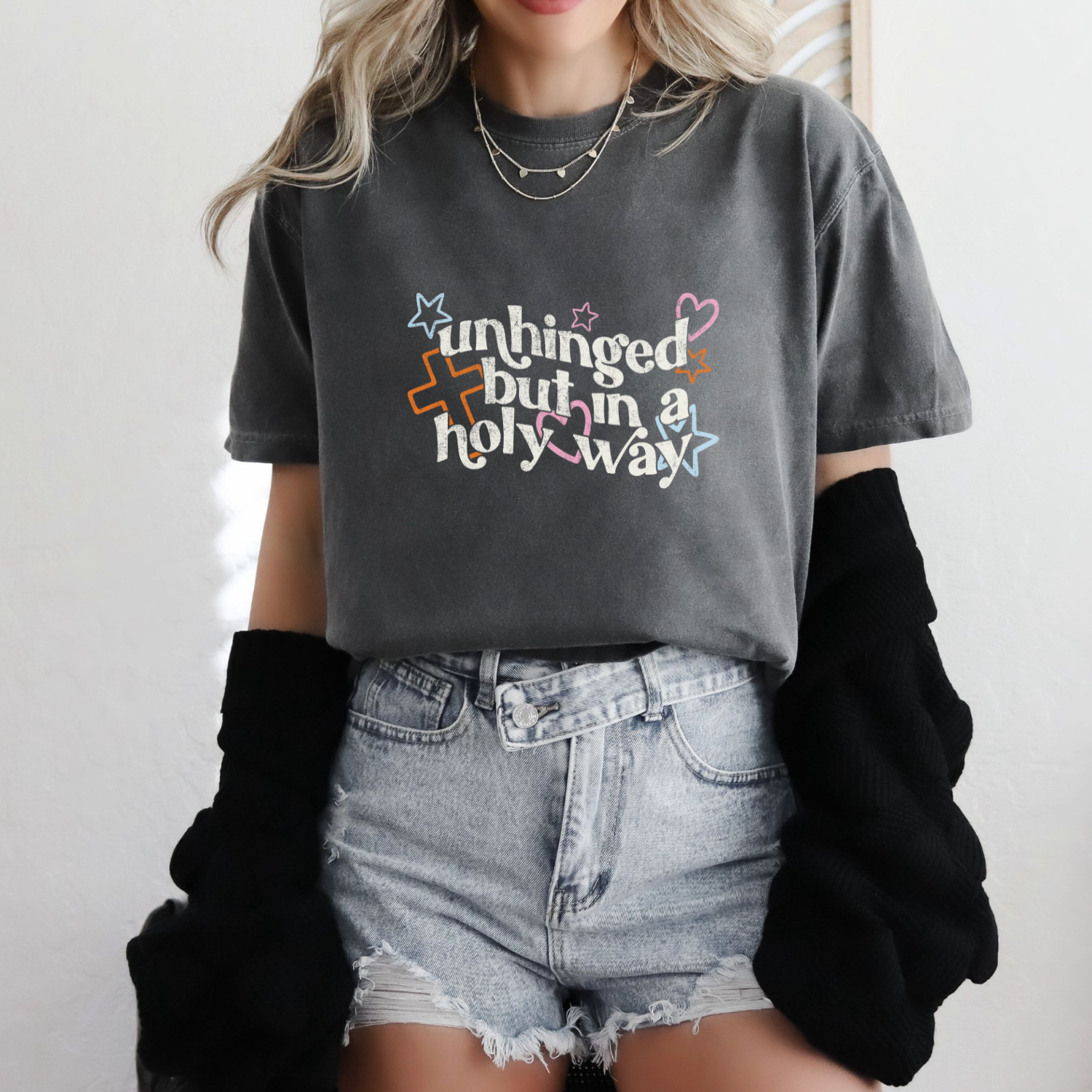 Person wearing a gray t-shirt with colorful text that reads "unhinged in a holy way", denim shorts, and black cardigan.