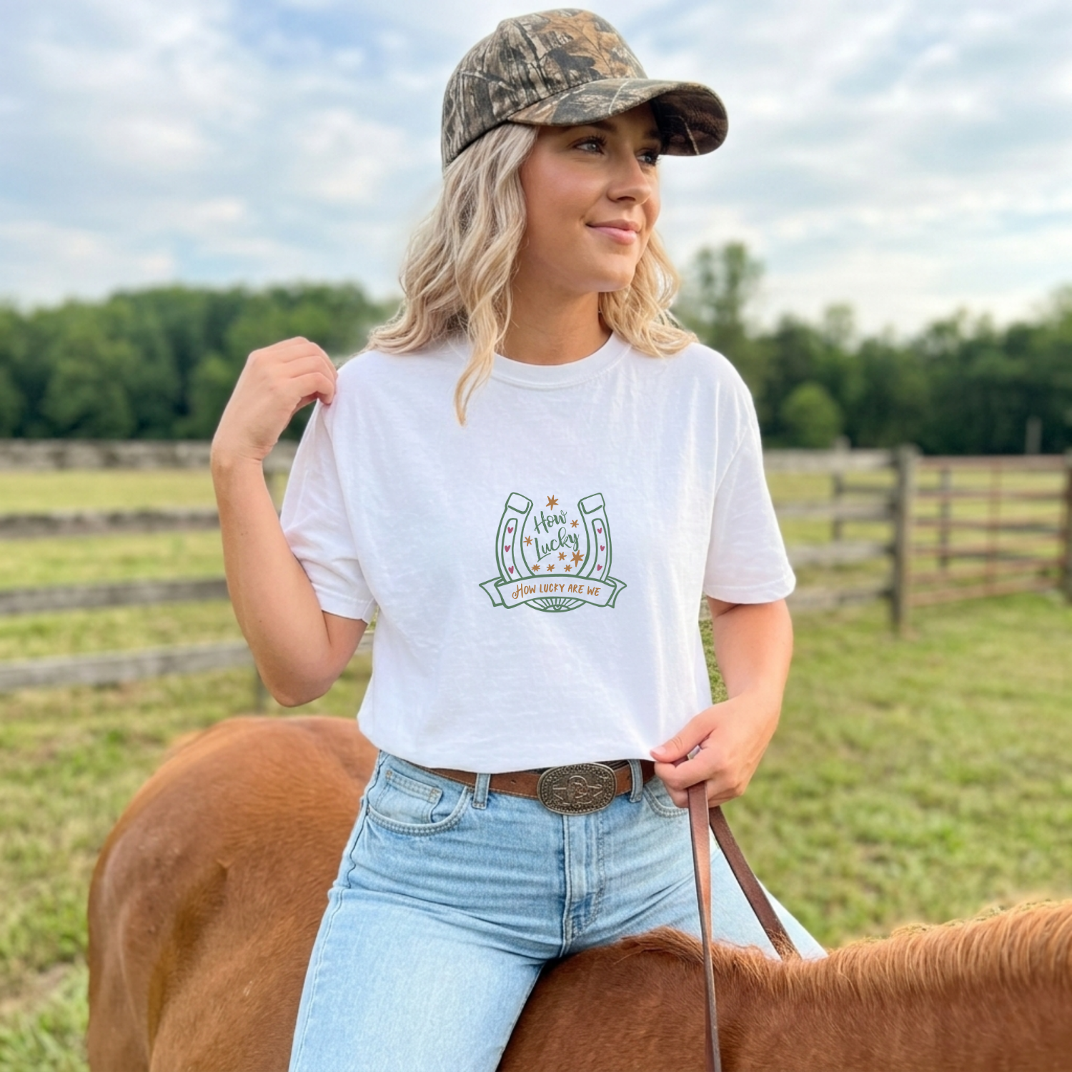 Woman in a white t-shirt with a cute horseshoe illustration, blue jeans, and camouflage cap standing next to a horse in a field.