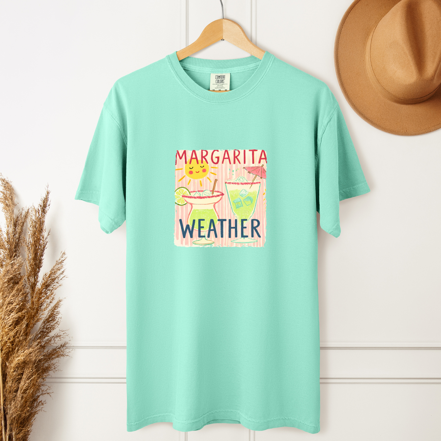 Mint green t-shirt with 'Margarita Weather' graphic on a wooden hanger against a white wall.