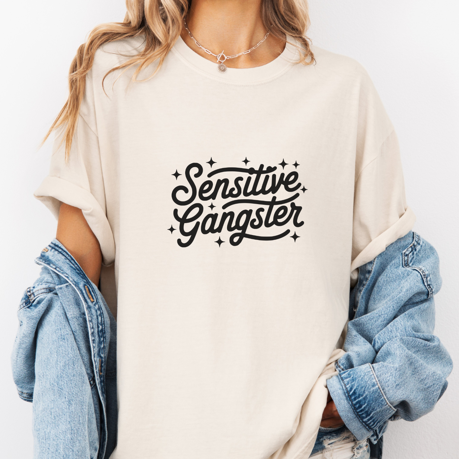 Person wearing a beige t-shirt with 'Sensitive Gangster' text, holding a denim jacket.