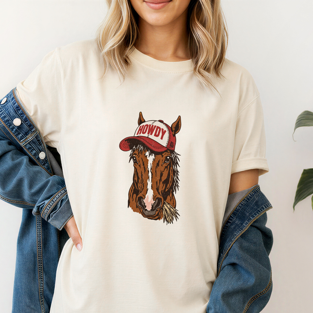 Person wearing a cream t-shirt with a horse graphic and 'Howdy' hat design, standing against a plain background.