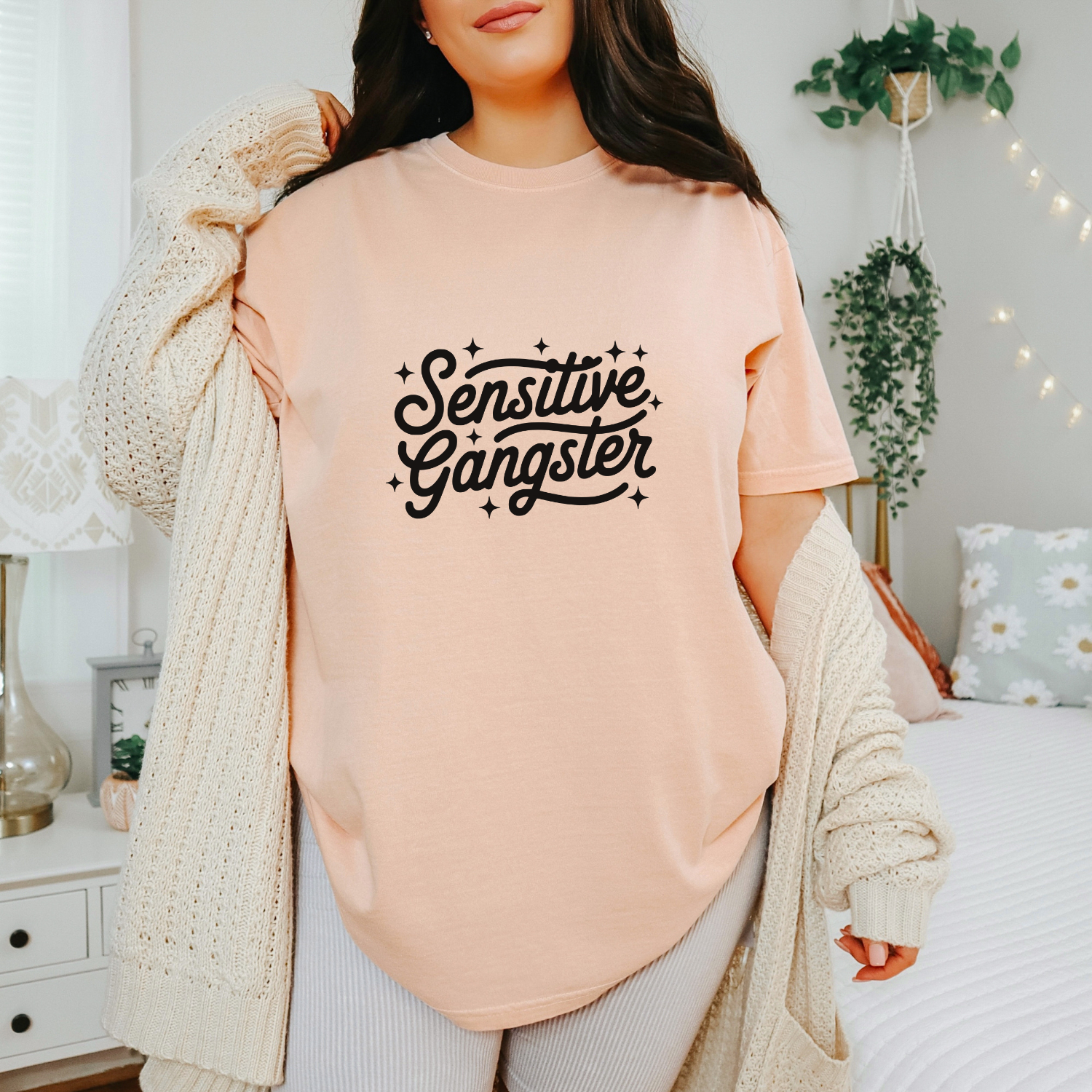 Person wearing a peach sweatshirt with 'Sensitive Gangster' text in a cozy room.