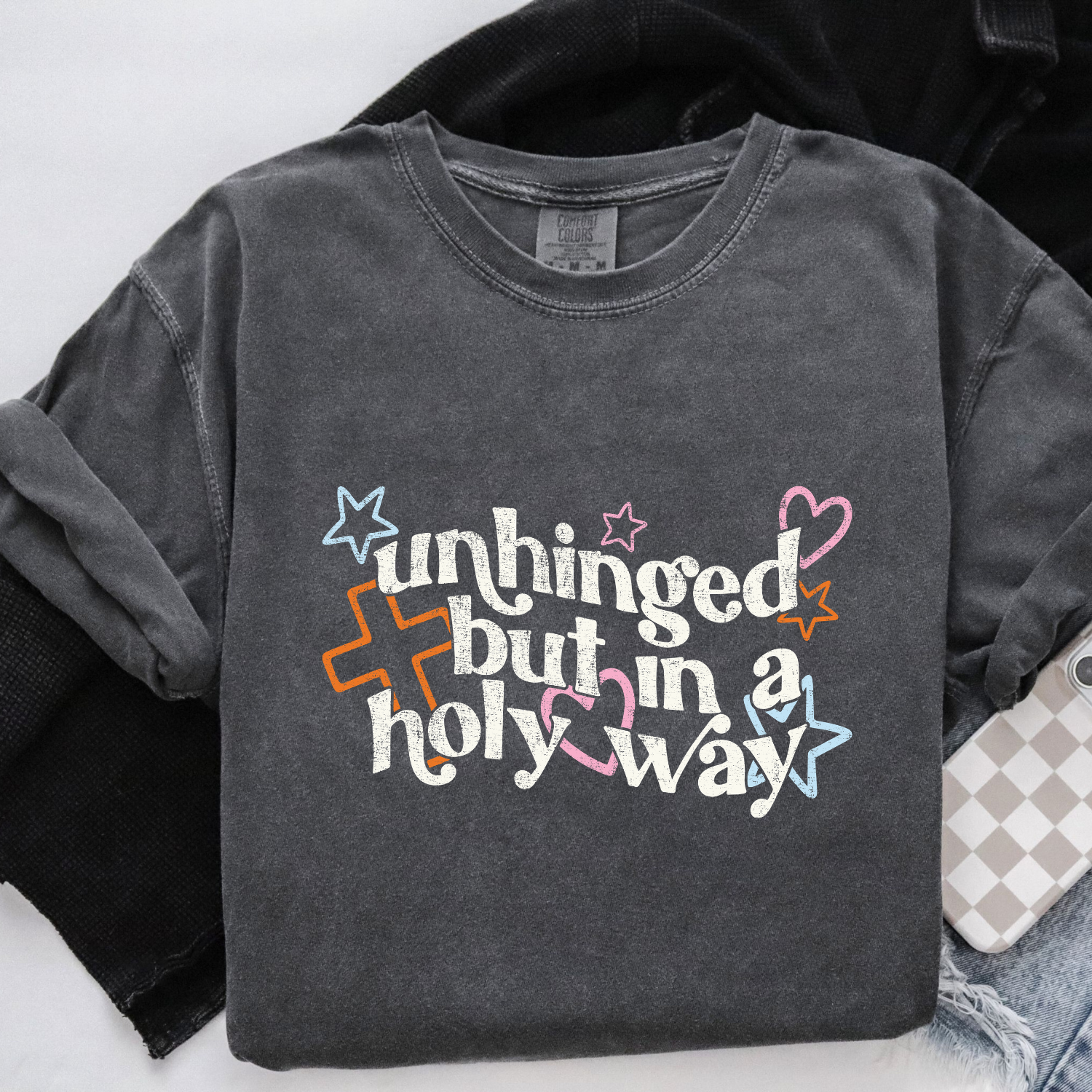 Dark gray sweatshirt with colorful text and graphics on a white background. The text reads "unhinged but in a holy way"