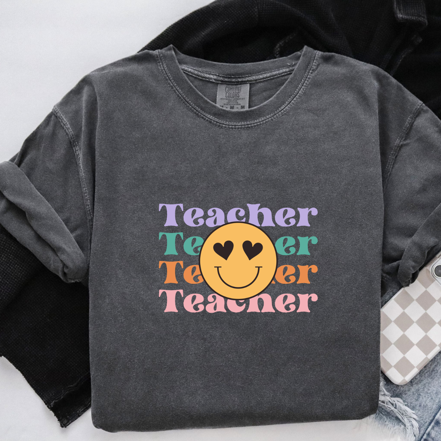 Vintage pepper colored comfort colors tee with a retro smiley face with hearts for eyes and the words "teacher" in the background in bright colors