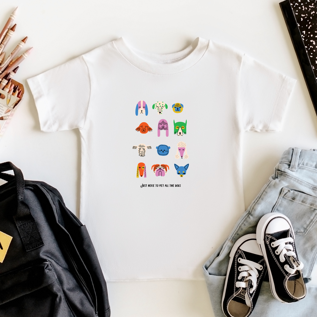 White t-shirt with colorful graphic design of dogs, black backpack, and sneakers on a white surface