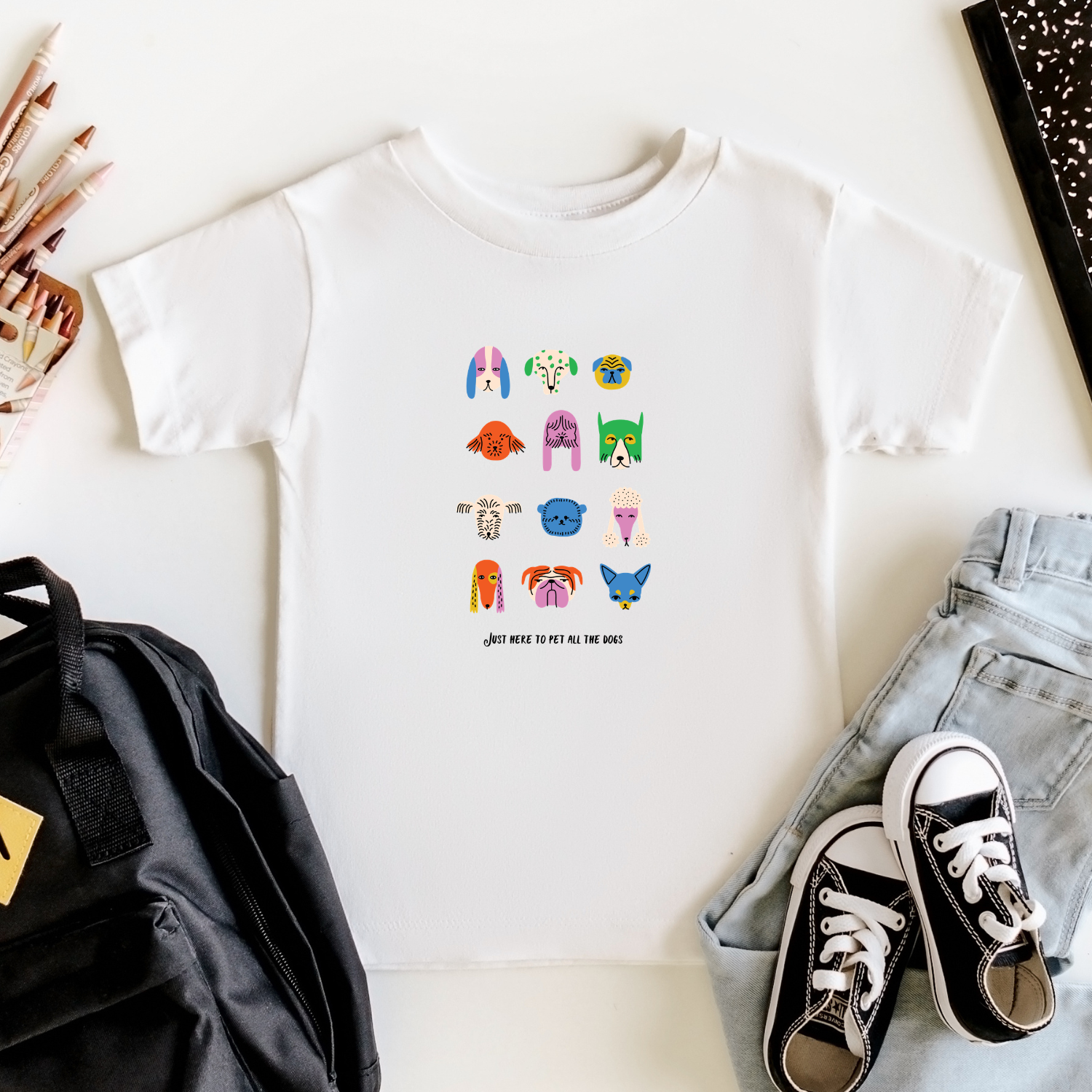 White t-shirt with colorful graphic design of dogs, black backpack, and sneakers on a white surface