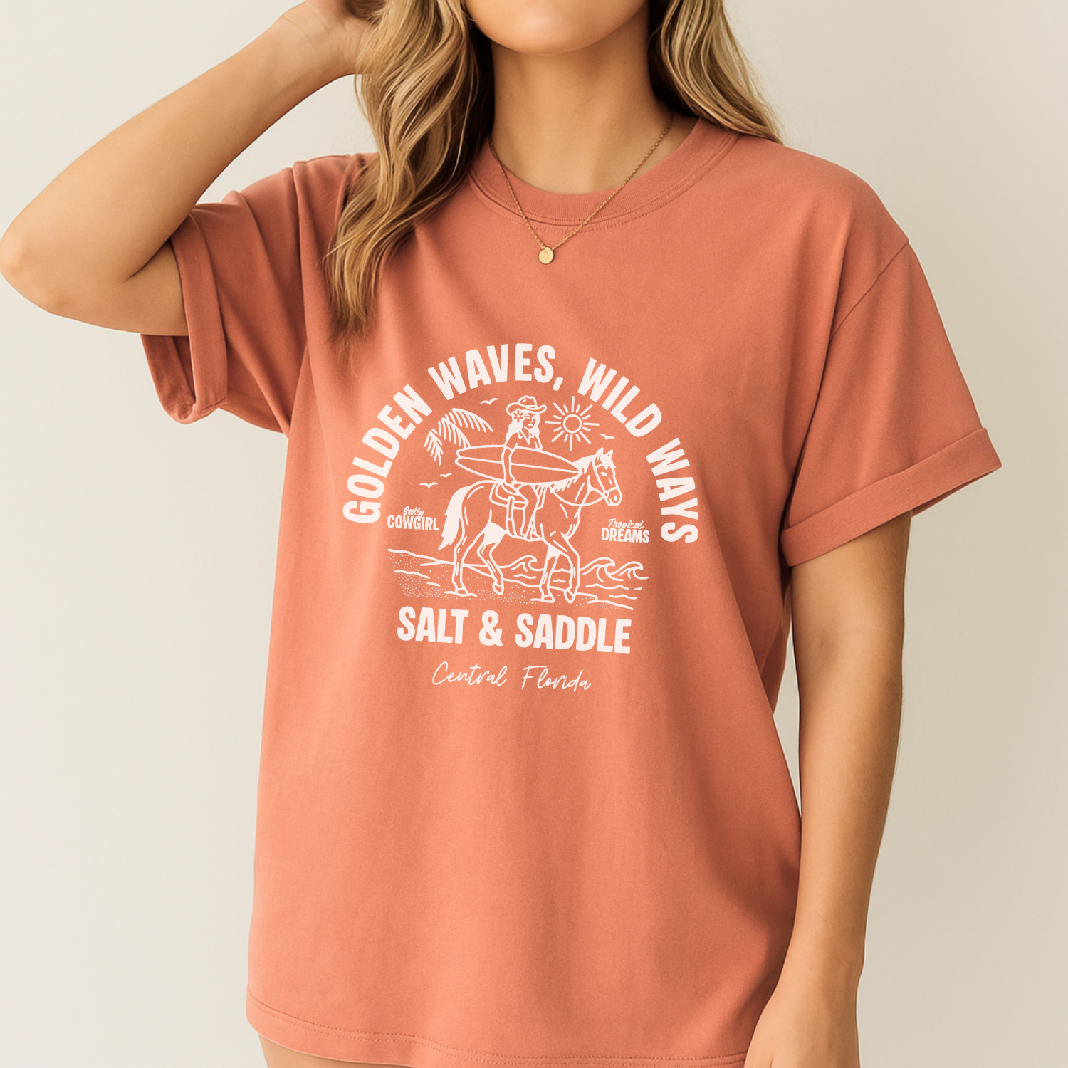 Terra Cotta comfort colors t-shirt with t-shirt with a white graphic of a cowgirl riding a horse along the beach. The girl is holding a surfboard. The text says salt and saddle, Central Florida. Coastal Cowgirl theme.