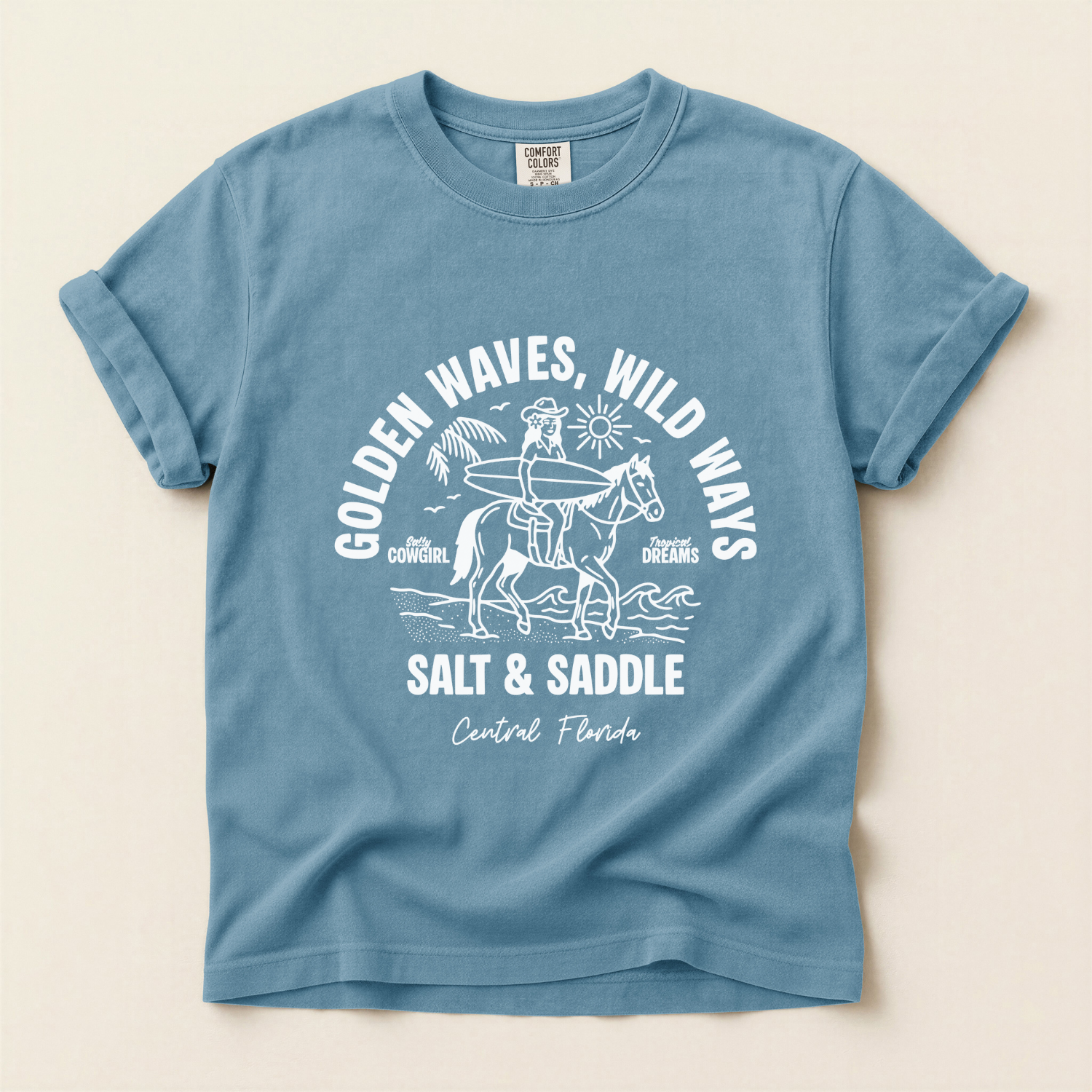 Blue comfort colors t-shirt with a white graphic of a cowgirl riding a horse along the beach. The girl is holding a surfboard. The text says salt and saddle, Central Florida. Coastal Cowgirl theme.
