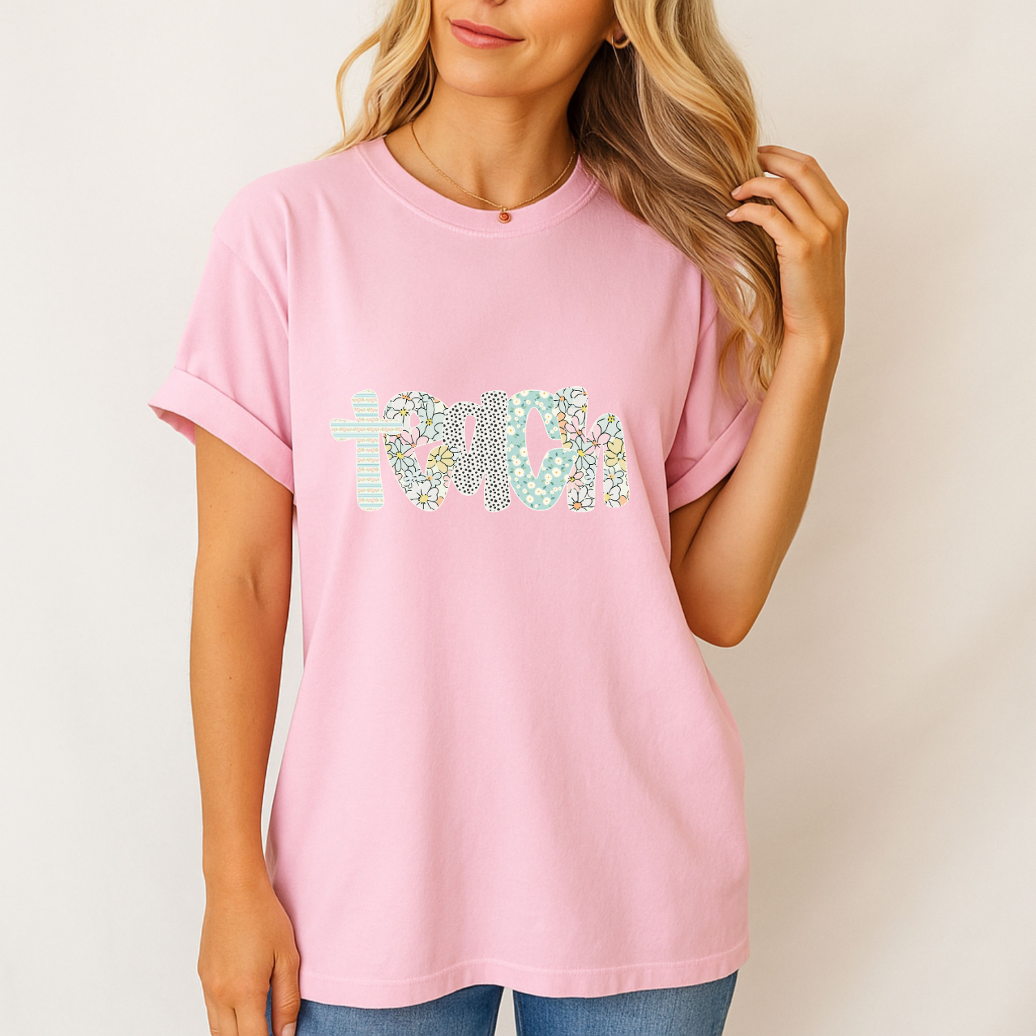 Person wearing a pink t-shirt with colorful text design on a plain background, floral lettering says teach